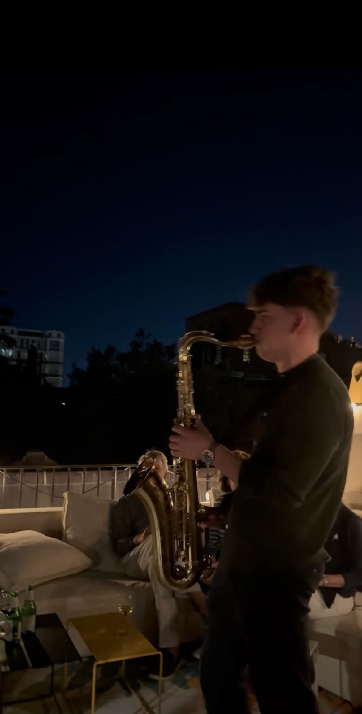 Jovan Pastar playing saxophone on a rooftop terrace after dark, guests on the lounge sofas beside him