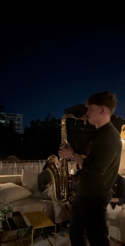 Jovan Pastar playing saxophone on a rooftop terrace after dark, guests on the lounge sofas beside him