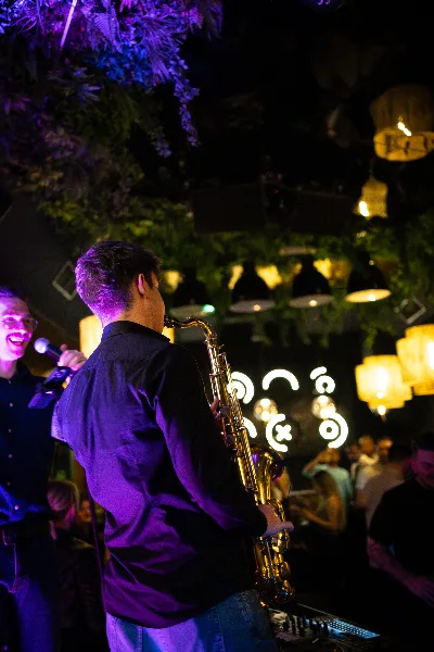 Jovan Pastar playing saxophone beside the DJ decks under purple light, lanterns over the crowd