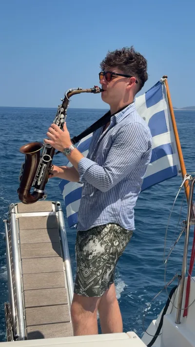 Jovan Pastar playing saxophone on the bow of a catamaran, a Greek flag in the wind behind him
