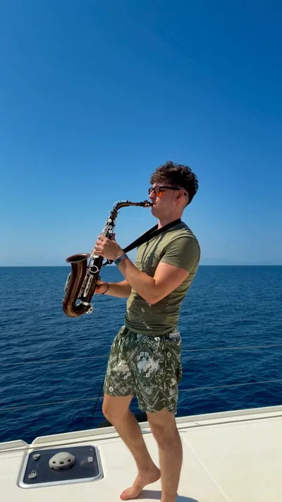 Jovan Pastar playing saxophone at the rail of a catamaran, the open sea behind him