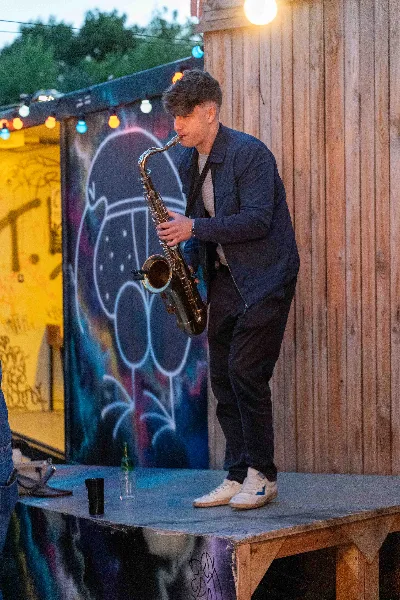 Jovan Pastar playing saxophone on a small open-air stage under string lights, street art behind him